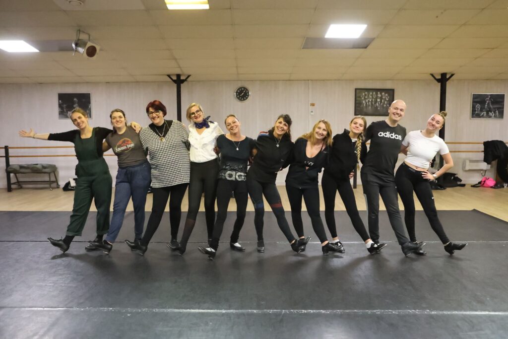 Groupe d’adultes en tenue de danse et chaussures de claquettes, posant bras dessus bras dessous dans un studio de danse, ambiance conviviale.