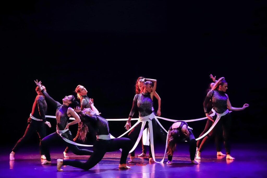 Groupe de danseurs sur scène, tous vêtus de tenues noires scintillantes avec des rubans blancs attachés à la taille. Chaque danseur adopte une pose expressive dans une chorégraphie contemporaine sous un éclairage violet