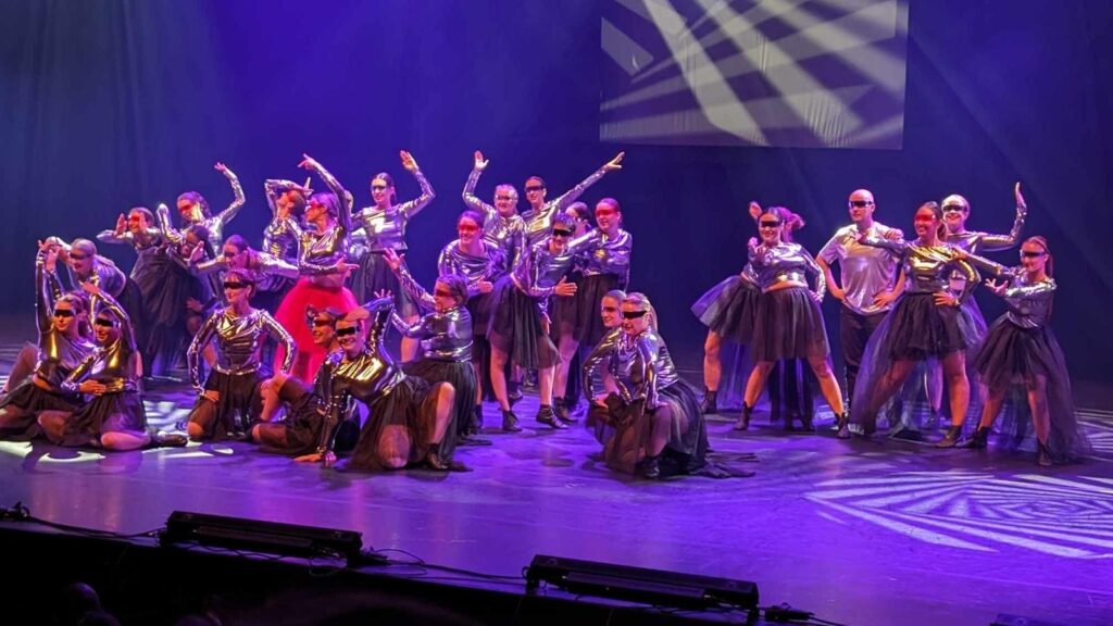 Grand groupe de danseurs et danseuses en costumes argentés et jupes noires, posant sur scène sous des lumières violettes lors d’un spectacle chorégraphié.