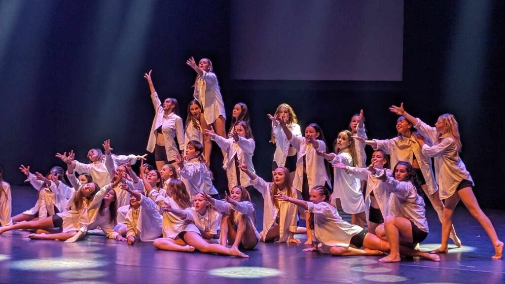 Ensemble de danseurs et danseuses vêtus de chemises blanches et shorts noirs, réalisant une pose expressive sur scène avec des éclairages artistiques.
