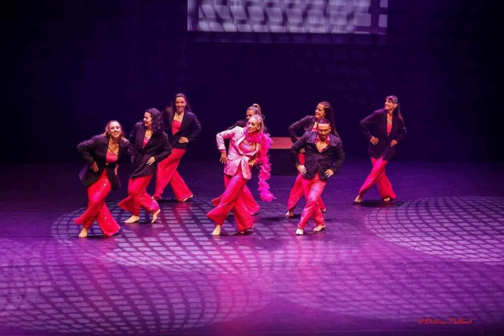 Troupe de danseurs en pantalon rose brillant et veste noire, effectuant une chorégraphie dynamique et souriante sur une scène éclairée de motifs lumineux violets, ambiance festive.