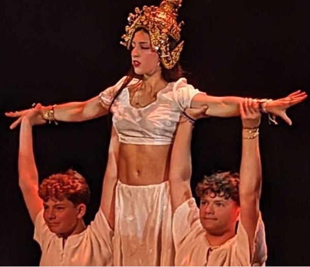 Jeune femme costumée, coiffée d’une couronne dorée, portée par deux garçons sur scène lors d’un spectacle de danse, fond noir et éclairage théâtral.